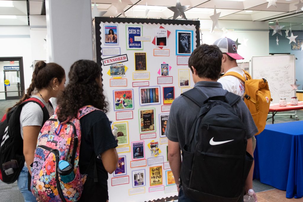 A group of four students observe a Banned Books Week display. The display features pictures of book covers and information on why they have been censored.