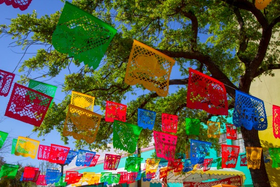 Rows of papel picado art hanging outside with trees and a blue sky