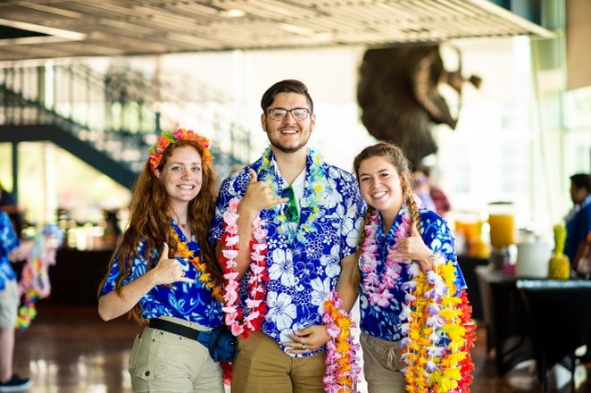 Three smiling students wearing floral print shirts and making hang-loose symbols with their hands.